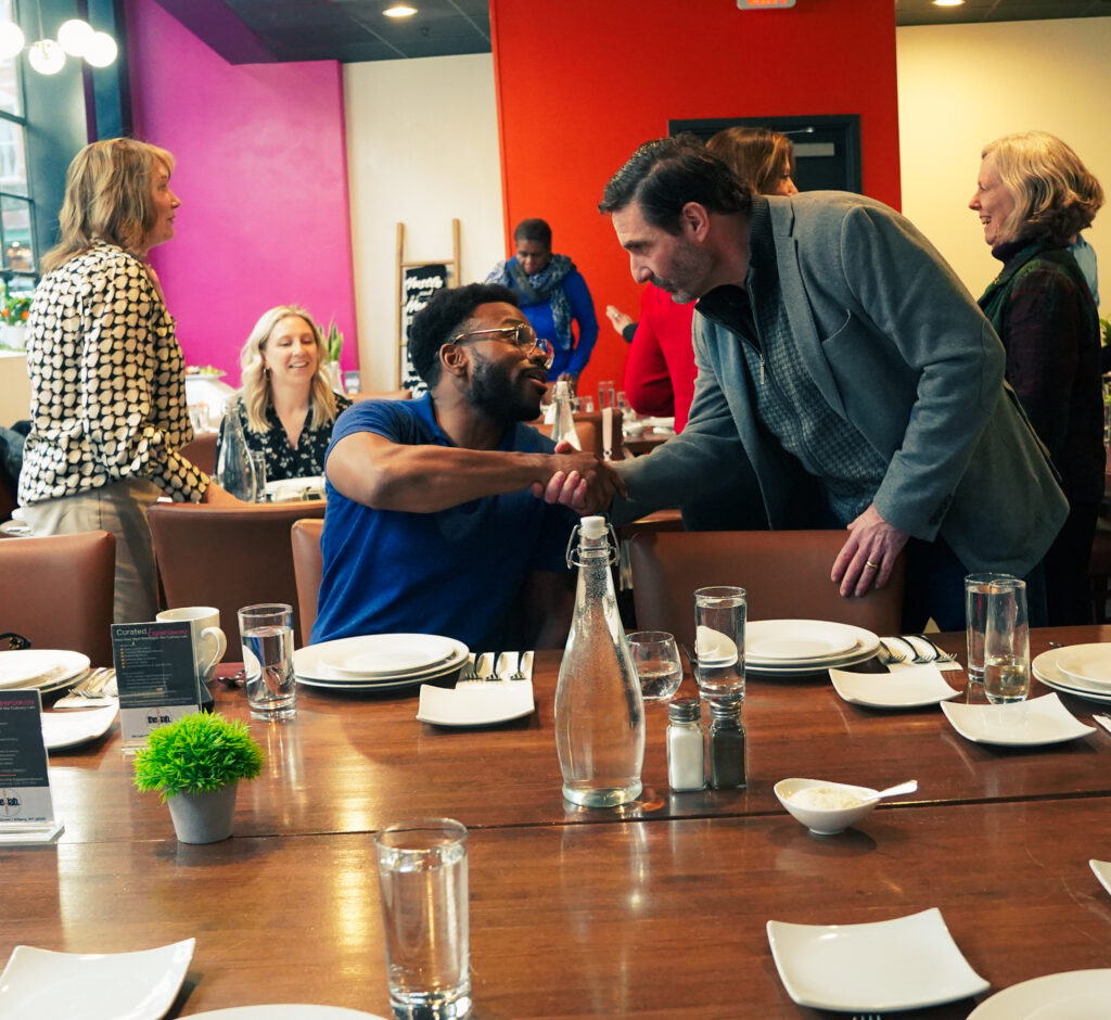 One Man Seated Shaking Hands with Another Man
